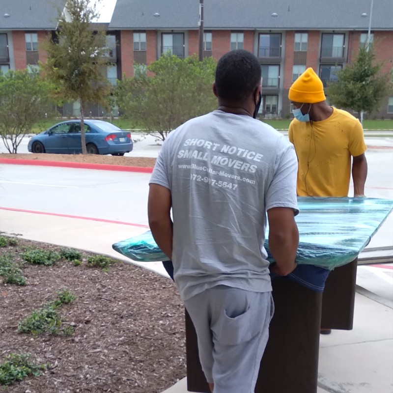Professional apartment movers in Fort Worth carefully transporting shrink-wrapped furniture at a residential complex.